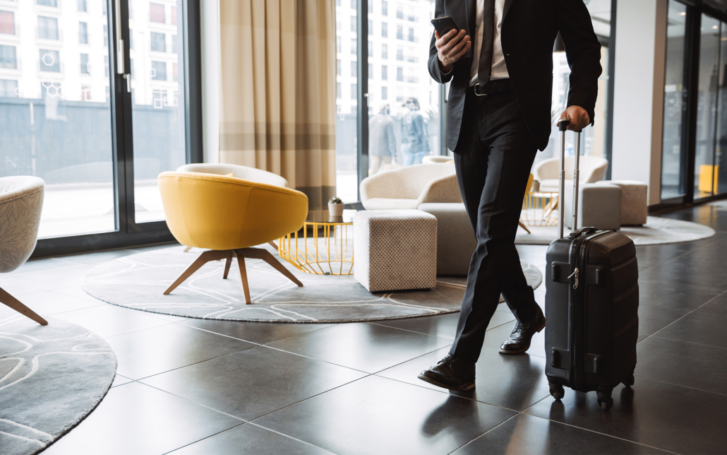 Cropped photo of successful businessman wearing suit holding smartphone and walking with suitcase in hotel lobby